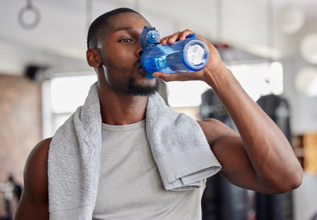 Young black male drinking water in the gym after workout with towel around neck
